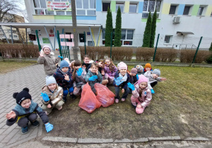 Zdjęcie przedstawia dzieci podczas akcji sprzątania terenu wokół przedszkola